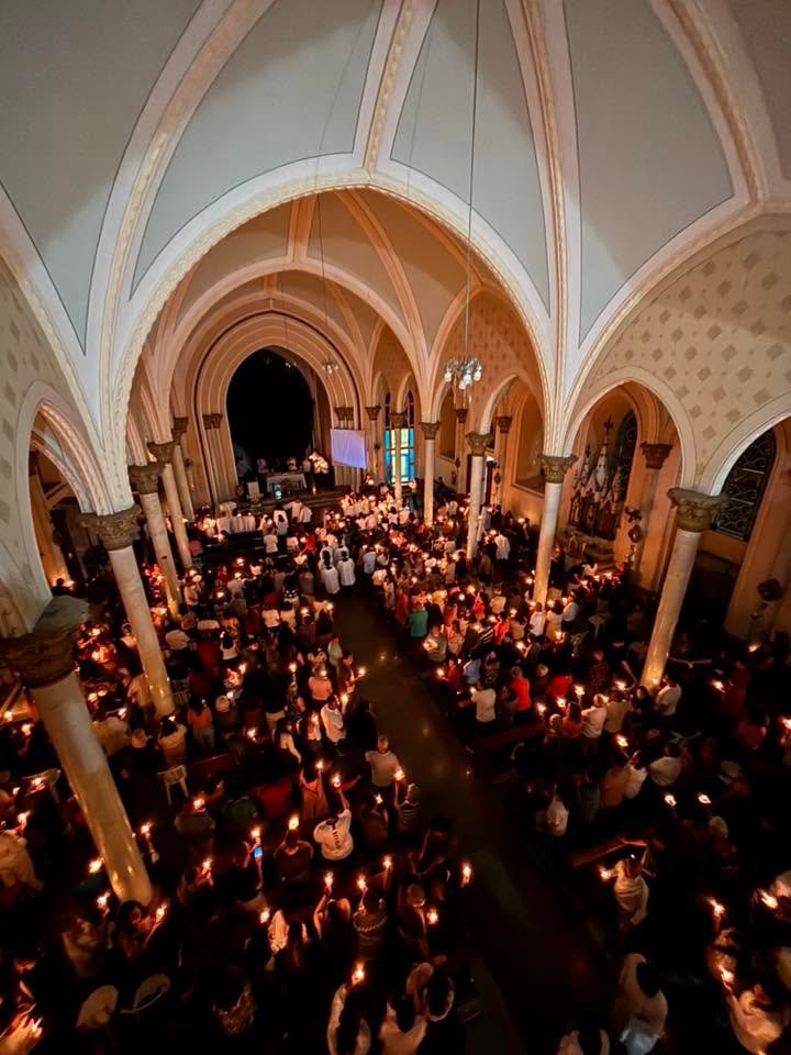 Fiéis com velas acesas lotando a nave da Igreja Matriz de Brazópolis, vista do coro durante celebração noturna