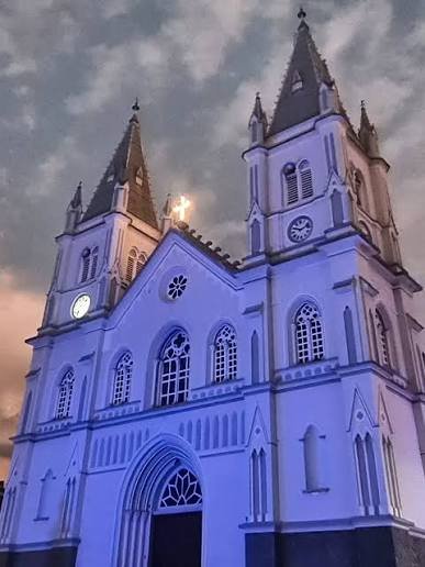 Igreja Matriz de São Caetano de Brazópolis iluminada à noite, com a fachada branca em tons de lilás e azul