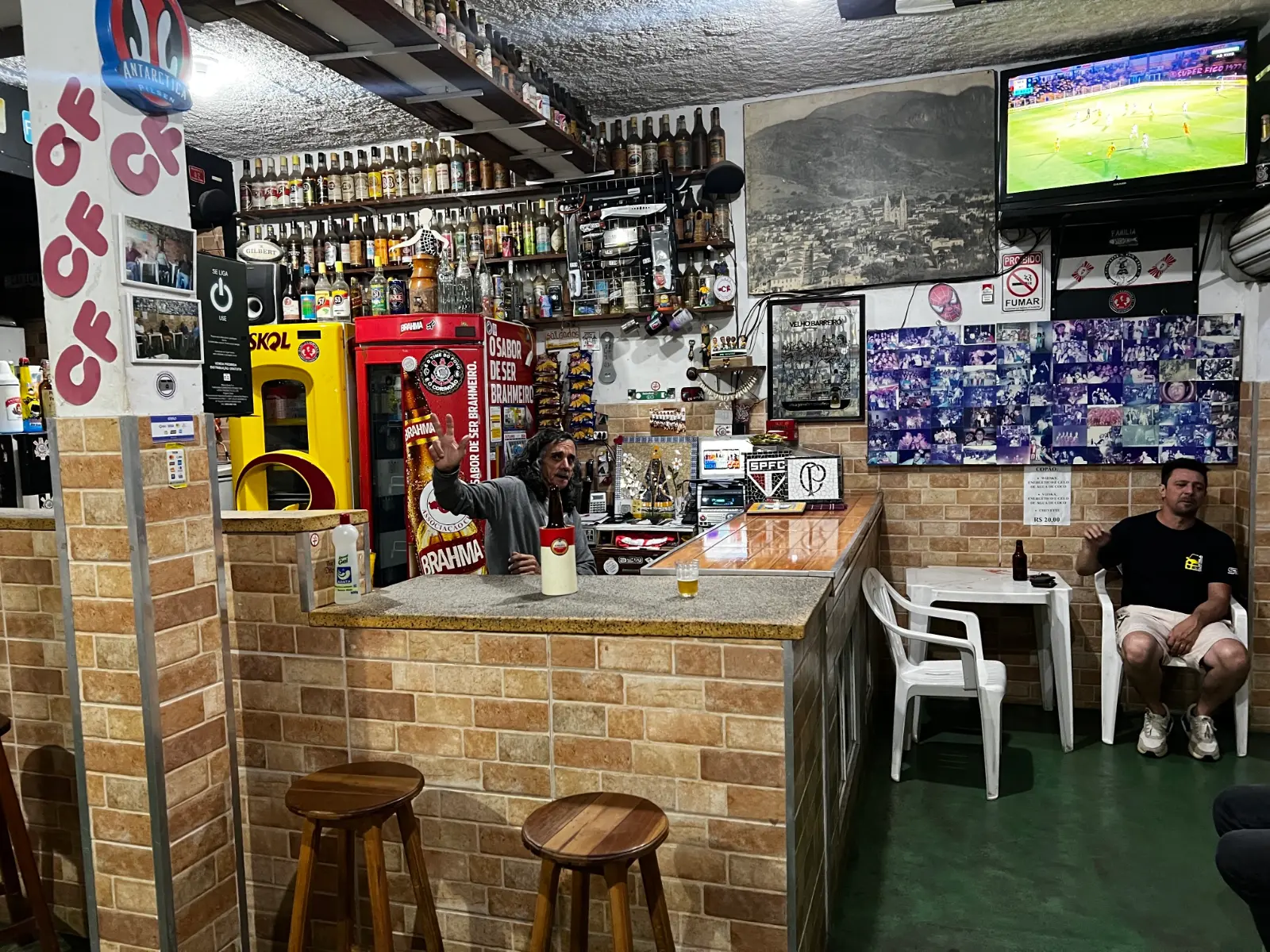 Balcão do Bar do Dito Cavichi com prateleiras cheias de garrafas de cachaça, foto antiga de Brazópolis e TV com futebol