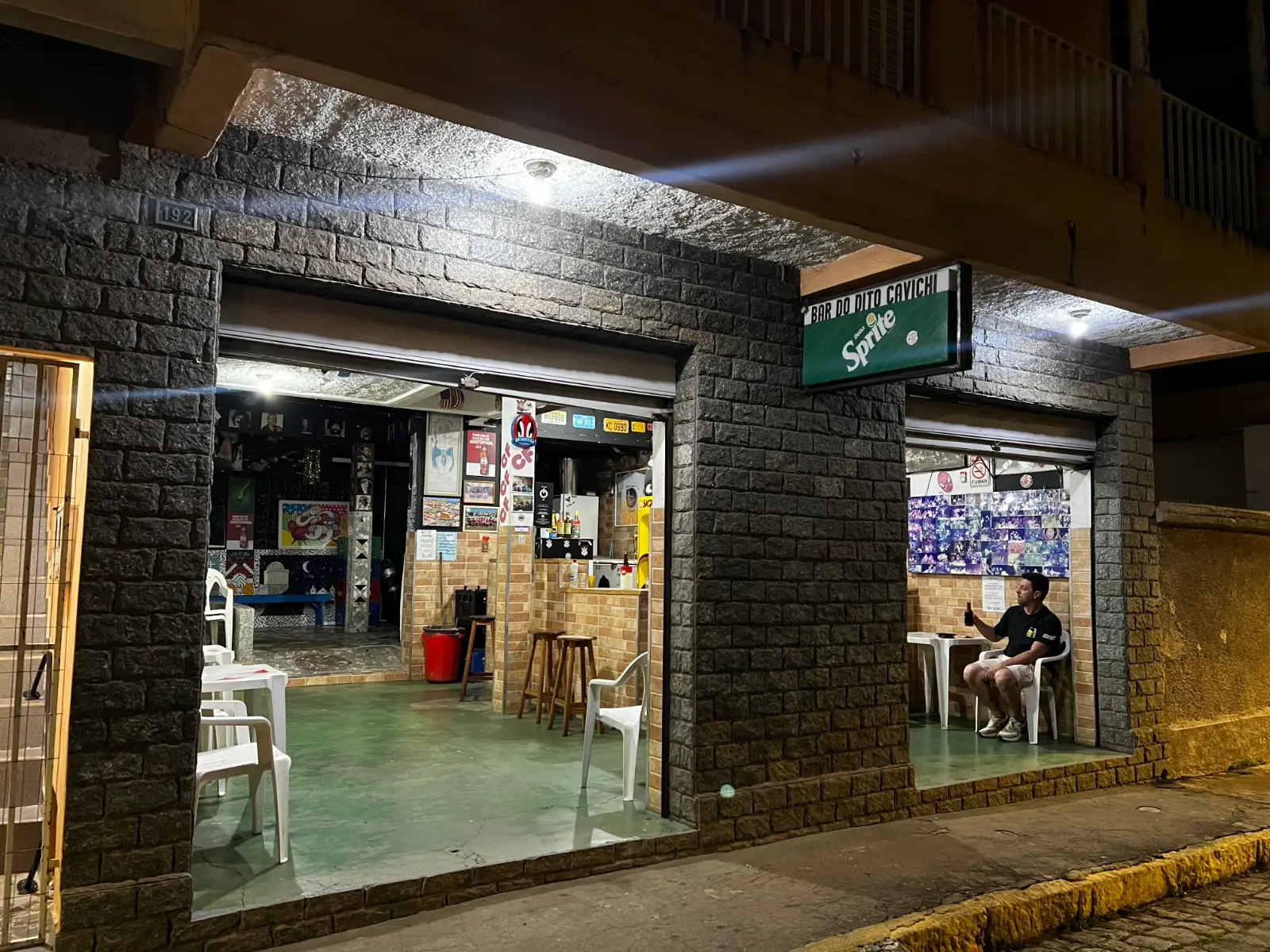 Bar do Dito Cavichi: boteco tradicional com música ao vivo em Brazópolis 11 Fachada do Bar do Dito Cavichi à noite, com paredes de pedra e portas abertas, na Rua José Pereira da Rosa, centro de Brazópolis MG