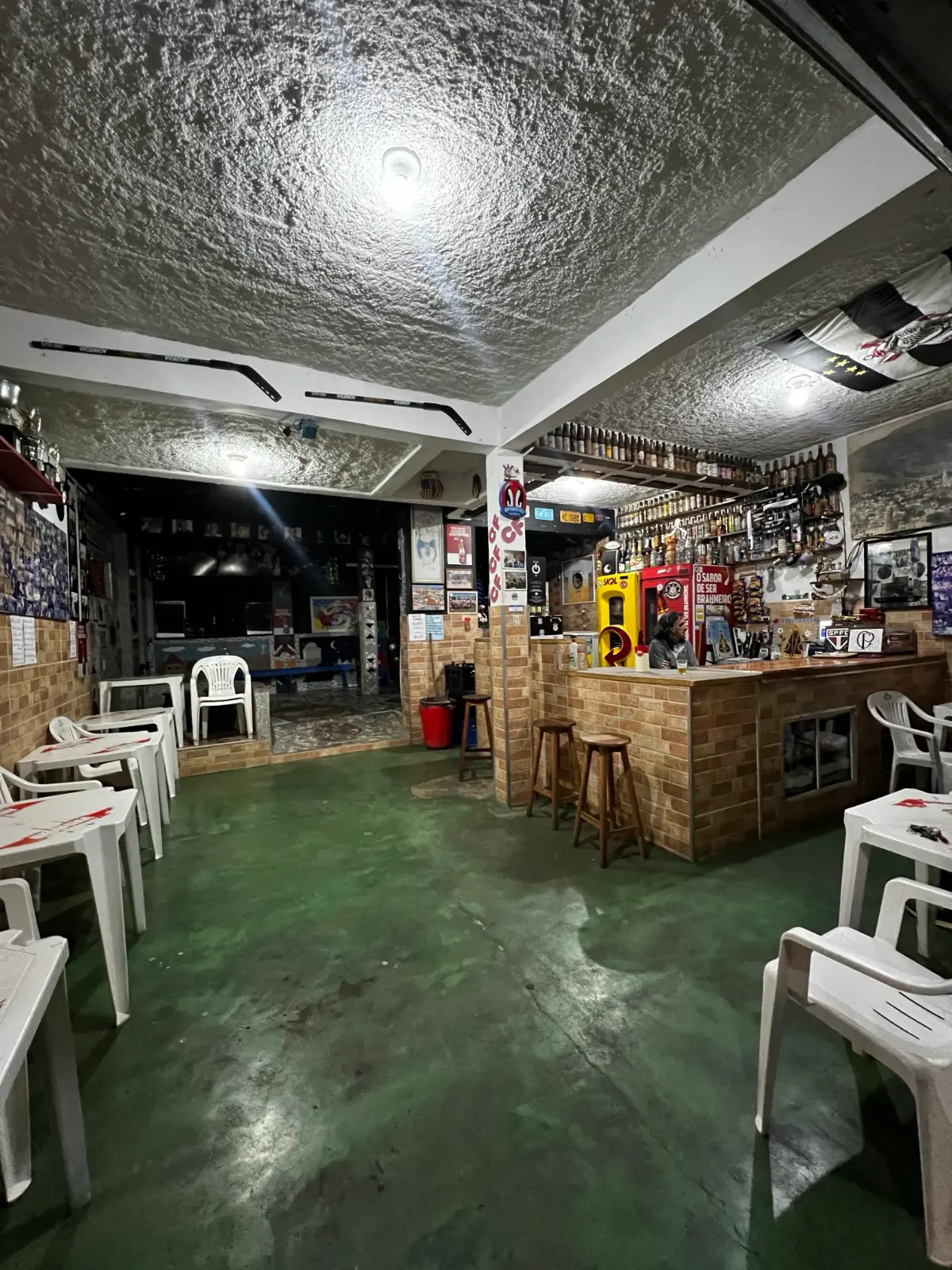 Salão do Bar do Dito Cavichi em Brazópolis com mesas, piso verde e balcão de tijolinho ao fundo
